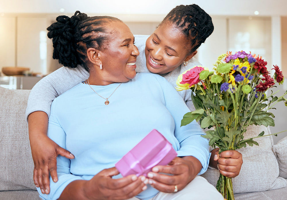 A woman holds a gift while another woman hugs her and presents a bouquet of colorful flowers, both smiling warmly in a cozy, well-lit living room.